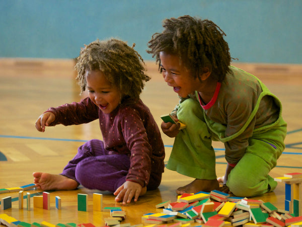 Zwei dunkelhäutige Kindergartenkinder spielen am Boden mit PESTAS Dominosteinen und haben richtig viel Spaß dabei.