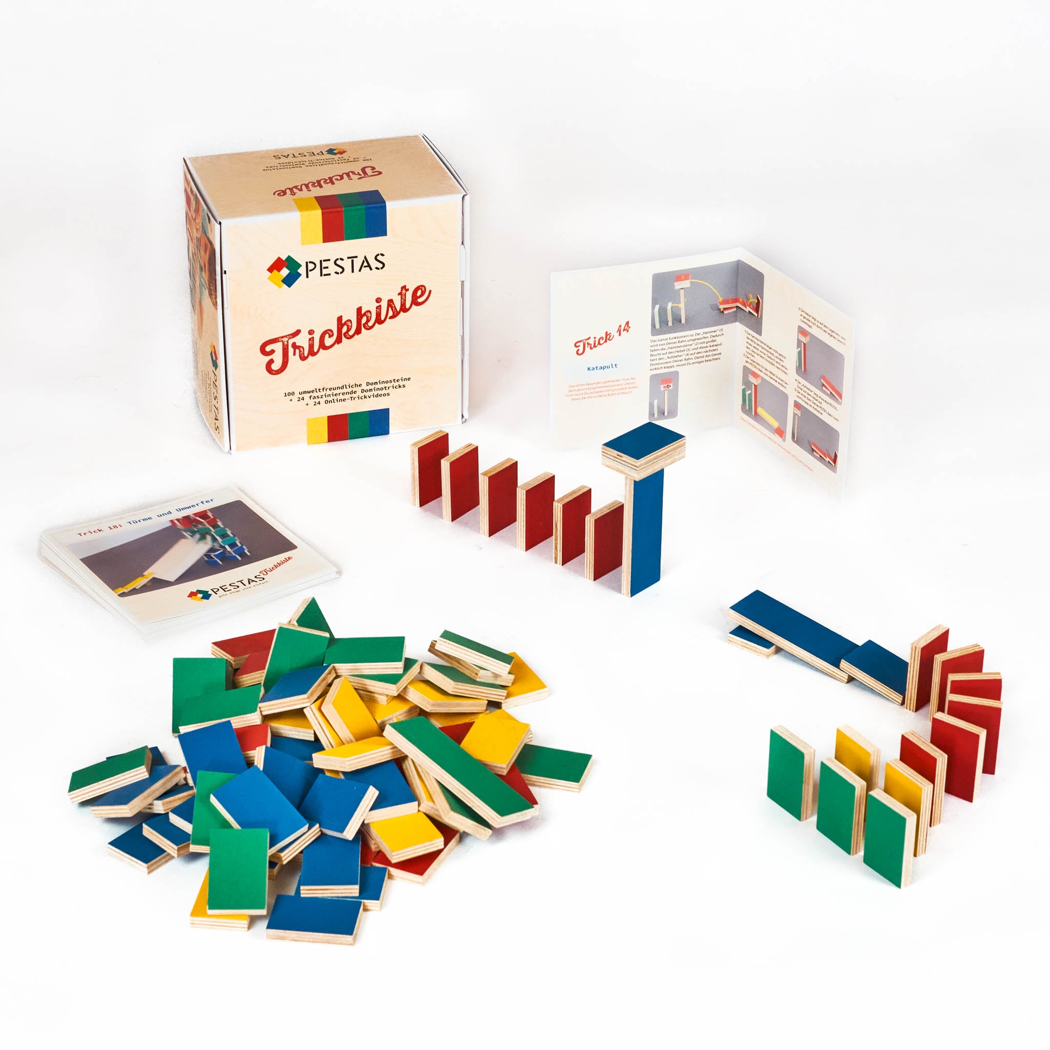 Produktbild PESTAS-Trickkiste | Das Bild zeigt die geschlossene PESTAS-Trickkiste. Davor ist der ganze Inhalt der Trickkiste aufgebaut: 24 Trickarten auf einem Stapel, eine davon aufgestellt. Der dazugehörige Trick (Trick 14, Katapult) ist mit PESTAS aufgebaut, die restlichen PESTAS-Steine der Packung liegen aufgehäuft davor.